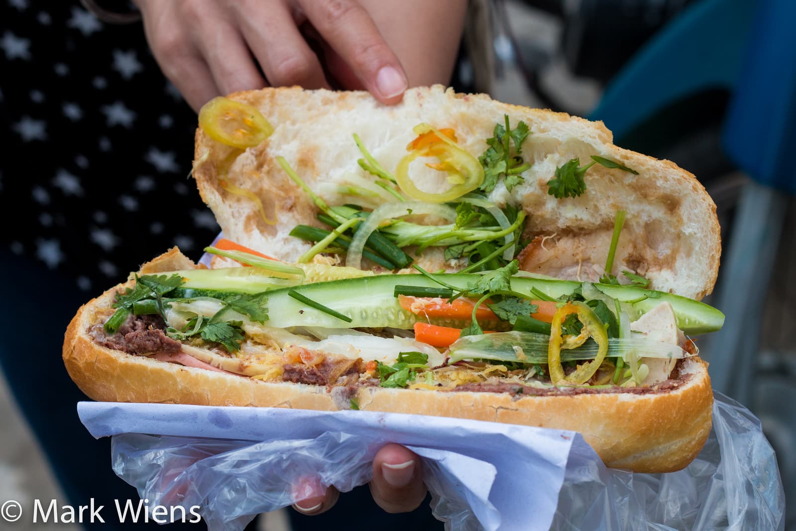 Street vendor filling a bánh mì baguette with meat and herbs at a cart in Hanoi