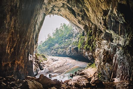 Tour group at the entrance of Hang En cave in Phong Nha, Vietnam, with sandy river beach and limestone cliffs.